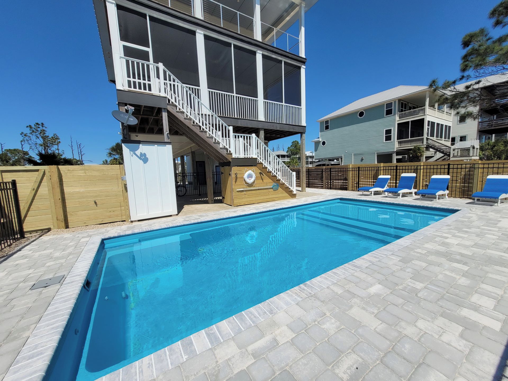 A swimming pool with a wooden deck, a covered area with a staircase, and a fenced area with lounge chairs.
