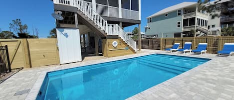 A swimming pool with a wooden deck, a covered area with a staircase, and a fenced area with lounge chairs.