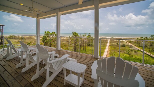 A covered deck with white Adirondack chairs and a small table, overlooking a beach and ocean. - Upscale Beach Front Home w/Elevator, Private Pool, Fire Pit! (Cape San Blas)