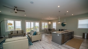 A spacious living room with a large window, a ceiling fan, and a dining area with a view of the outdoors. - Upscale Beach Front Home w/Elevator, Private Pool, Fire Pit! (Cape San Blas)