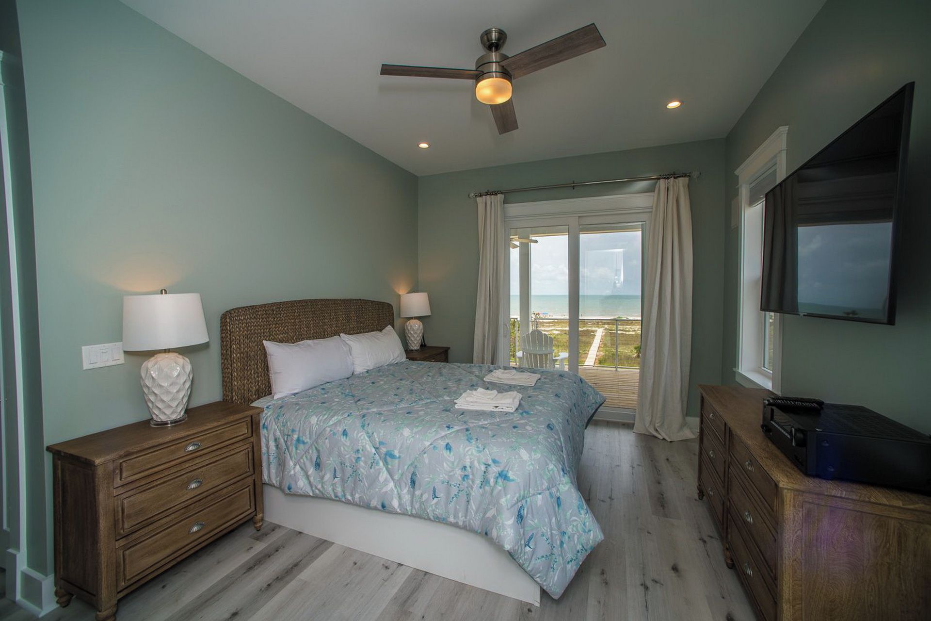 A bedroom with a bed, two bedside tables, a ceiling fan, a TV, and a view of the ocean.