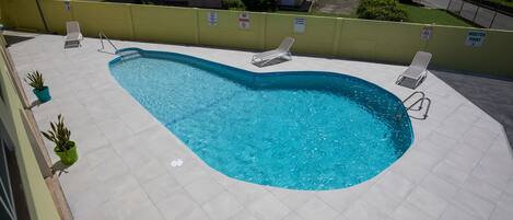 Una piscina con borde curvo, rodeada por un muro amarillo y una valla blanca. Hay sillas de descanso y plantas en macetas a lo largo de la piscina.