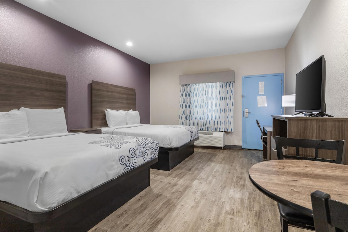 A hotel room with two beds, a dining table, a TV, and a bathroom with a shower.
