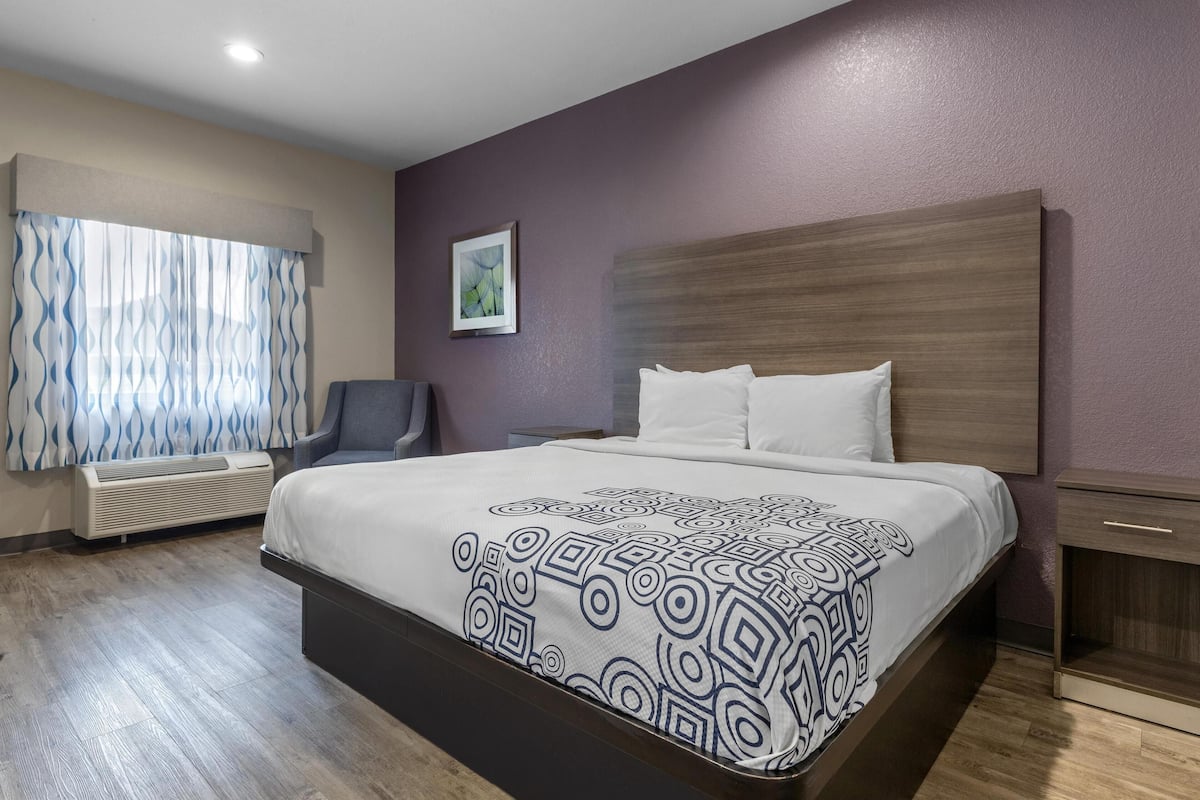 A hotel room with a large bed, a chair, a bedside table, and a wall-mounted air conditioner.