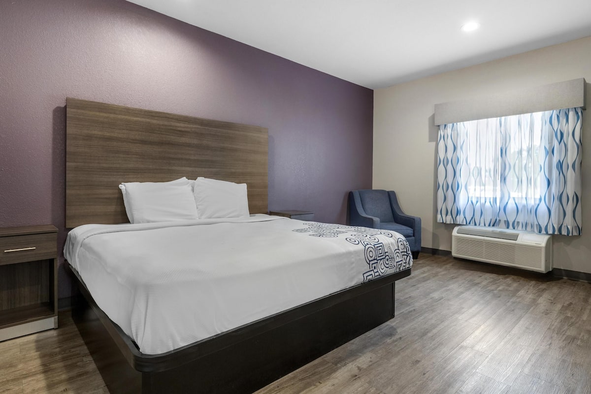 A hotel room with a large bed, a chair, a nightstand, and a window with curtains.