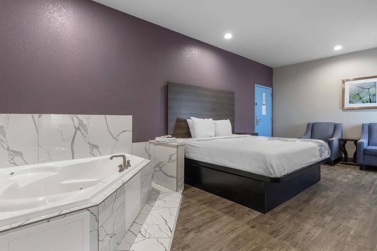 A modern bedroom with a large bed, a separate bathroom with a bathtub, and a small seating area.