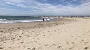 Une vaste plage de sable avec des empreintes, un ciel dégagé et des bâtiments qui se dessinent à l’horizon.