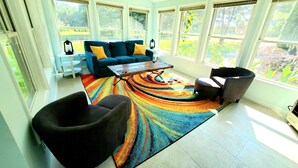 Un salón amplio con un sofá azul, una alfombra colorida y vistas a un jardín a través de grandes ventanales.