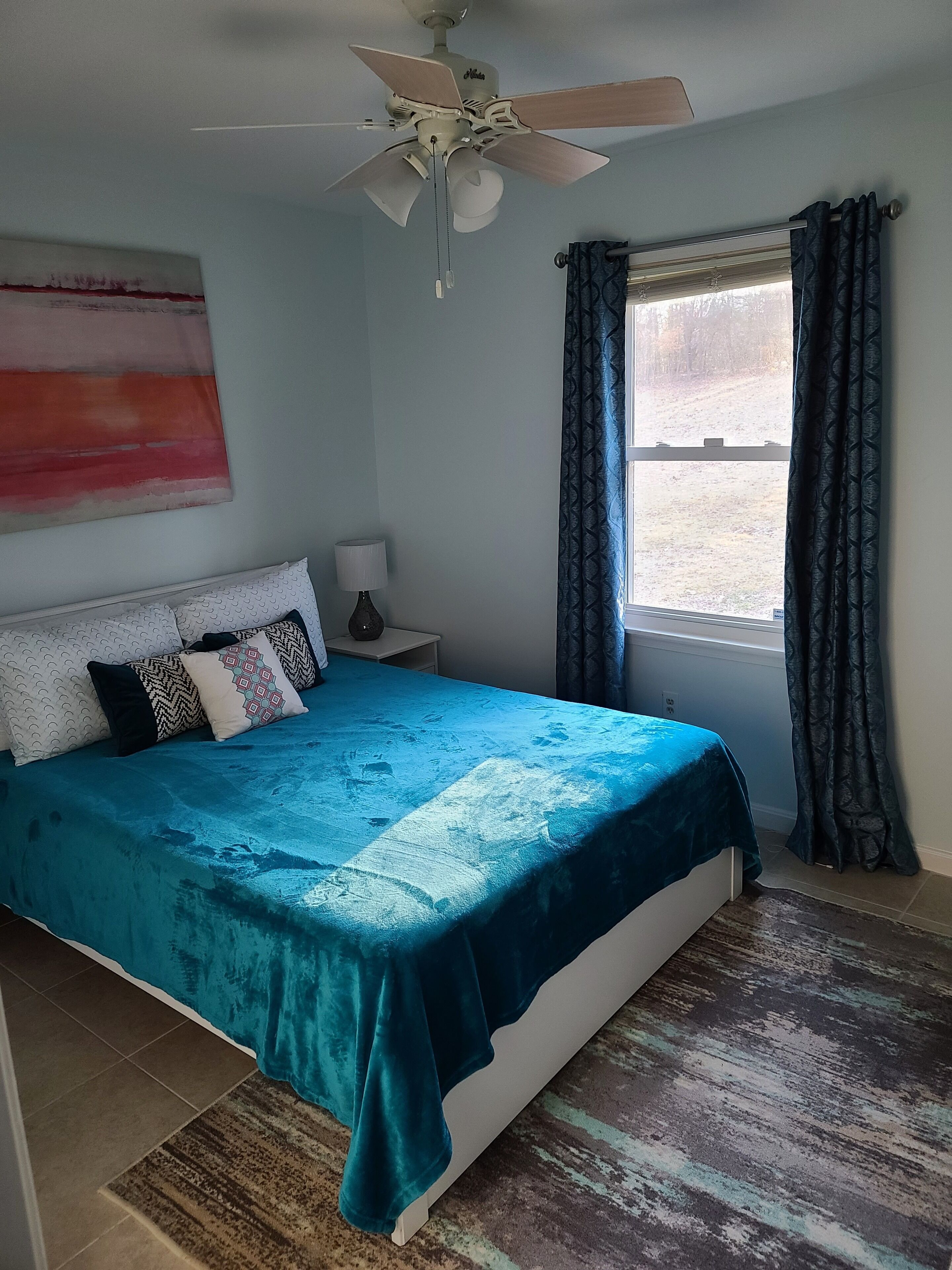 A bedroom with a bed, a ceiling fan, a window with curtains, and a wall painting.