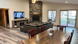 Een ruime woonkamer met een stenen openhaard, een grote modulaire bank, een houten eettafel met stoelen en een televisie op een houten dressoir.