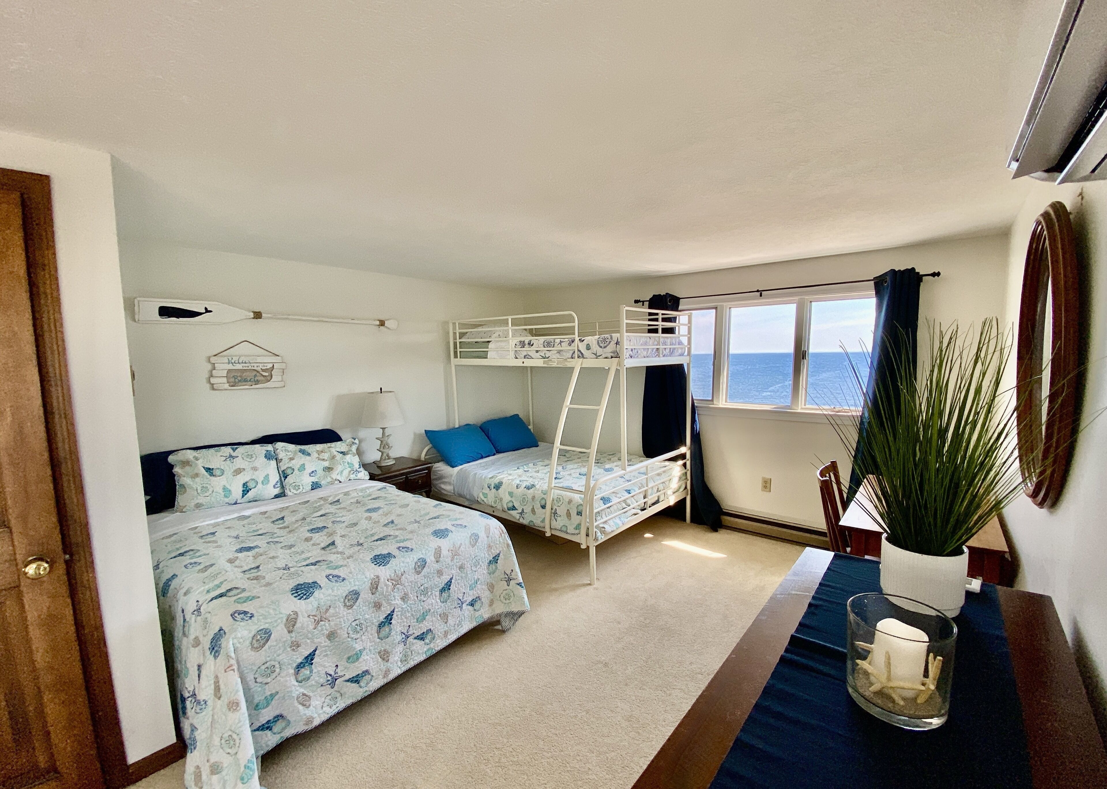A bedroom with two beds, a bunk bed, a window with a view, a potted plant, and a bedside table with a lamp.