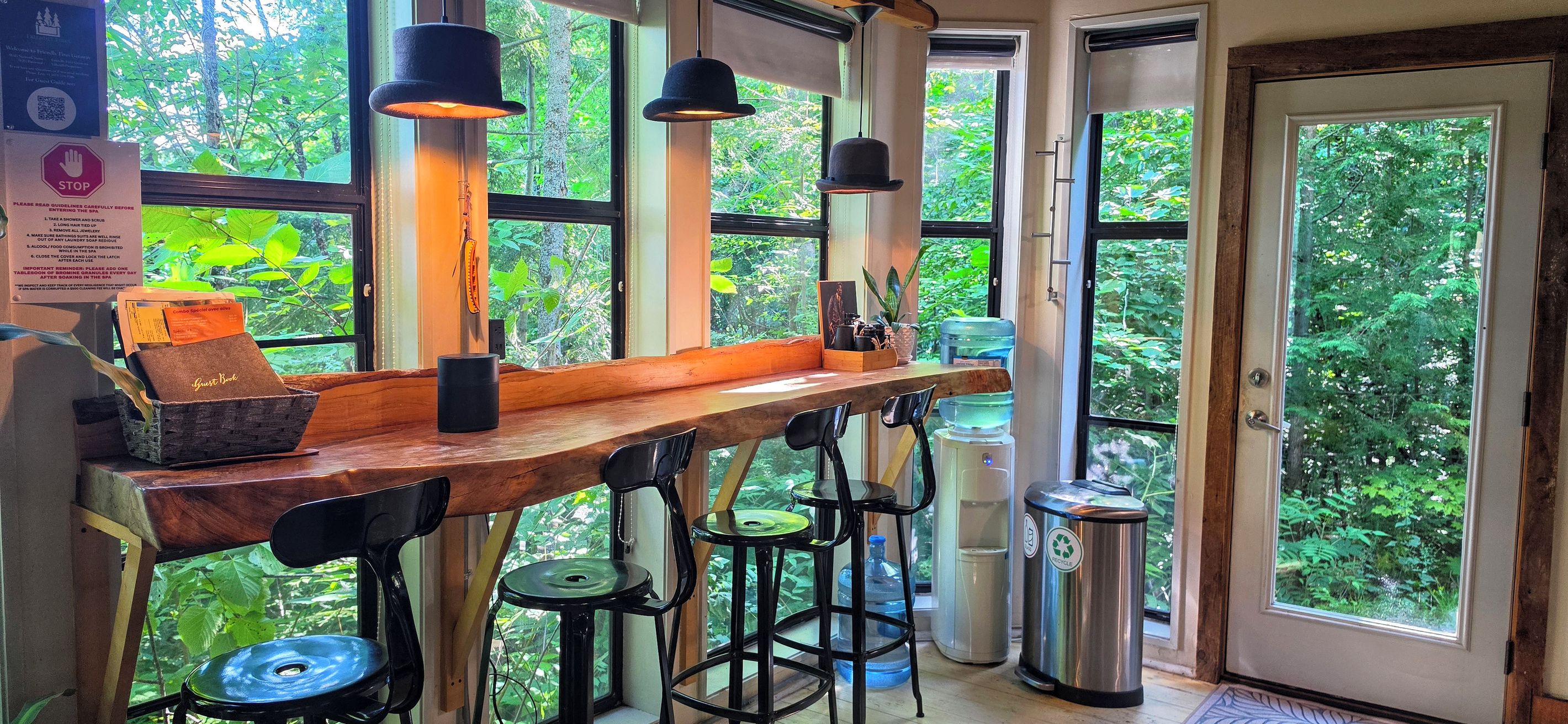 Un espace bar doté d’un comptoir en bois, de tabourets noirs, de luminaires suspendus et offrant une vue sur la verdure grâce à de grandes fenêtres.