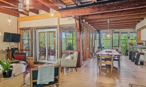 A spacious room with a wooden ceiling, large windows, a dining area with a table and chairs, and a television on the wall.