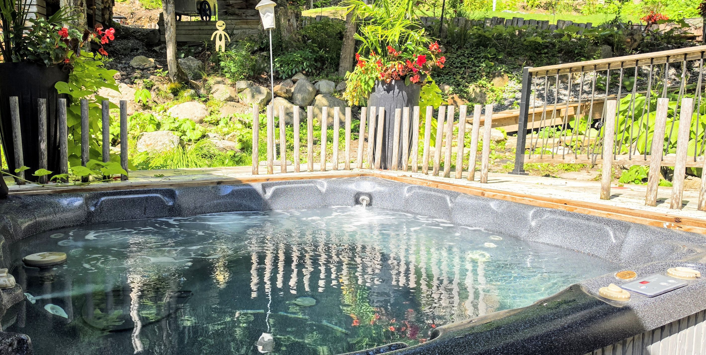 A hot tub surrounded by a wooden fence, with a garden and a wooden deck in the background.