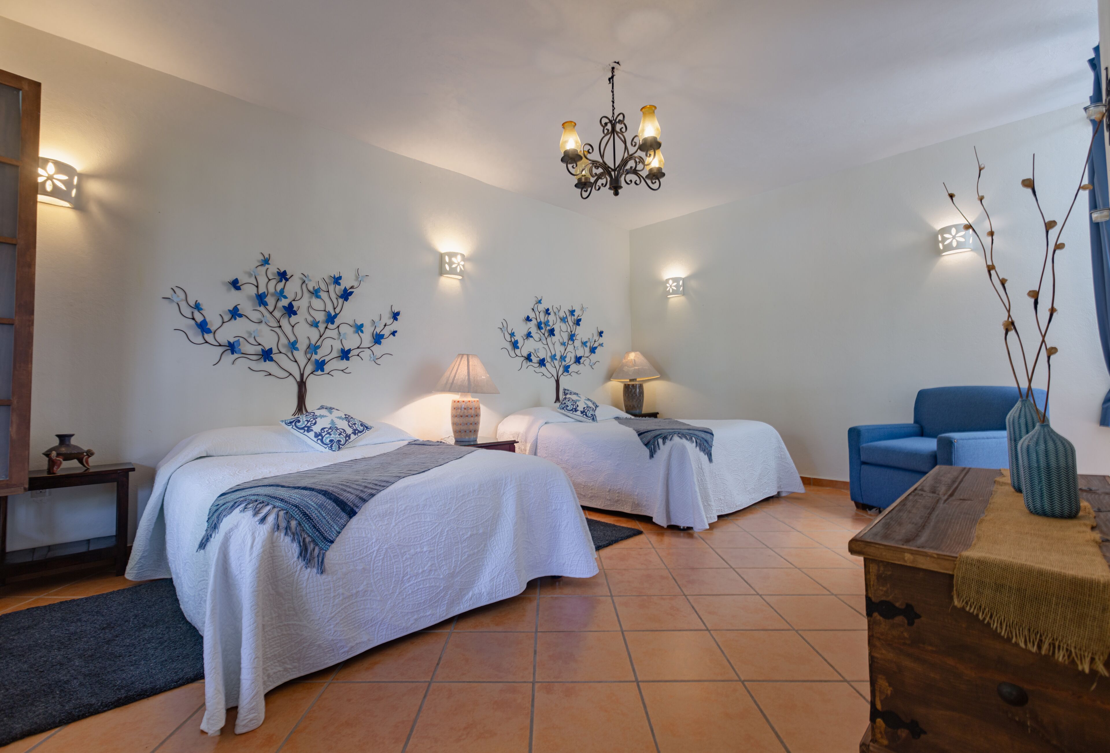 A room with two beds, a chandelier, wall sconces, and a blue armchair.