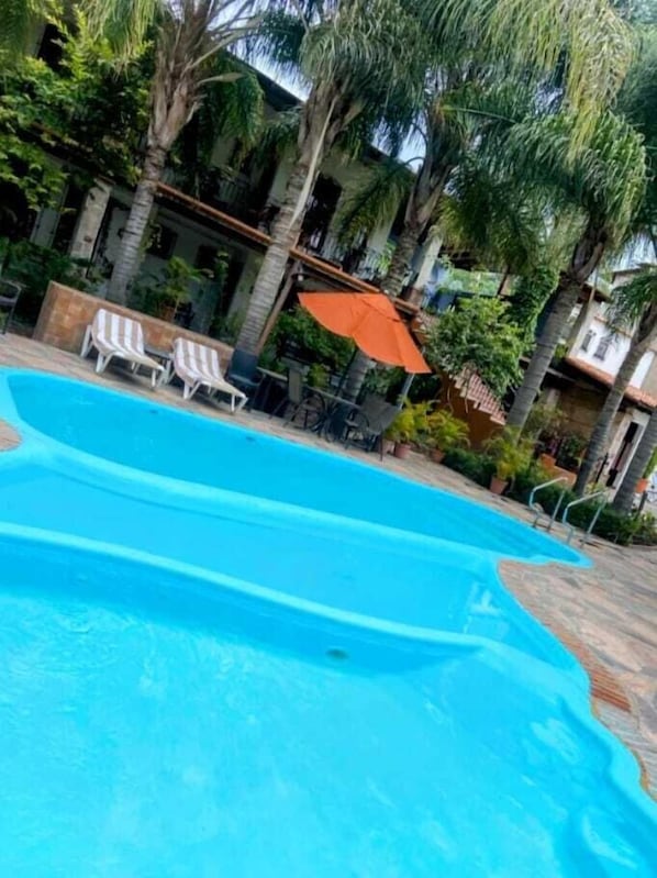 A swimming pool with lounge chairs and umbrellas, surrounded by palm trees and a building.