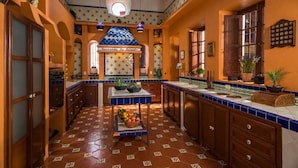 Une cuisine avec un sol carrelé, des meubles en bois, un îlot central orné de fruits et une enseigne décorative au-dessus de l’entrée.