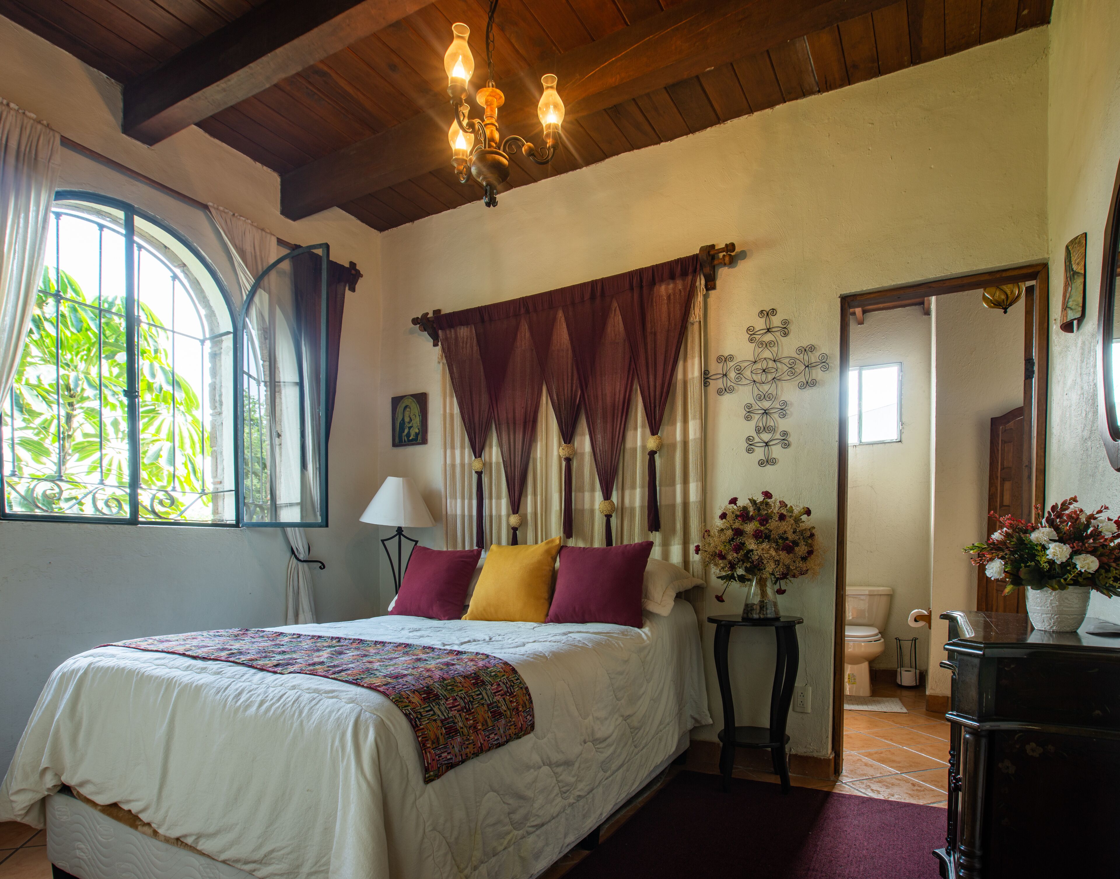 A bedroom with a large bed, a chandelier, a window with curtains, a bedside table with a lamp, and a mirror.