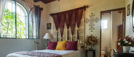 A bedroom with a large bed, a chandelier, a window with curtains, a bedside table with a lamp, and a mirror.