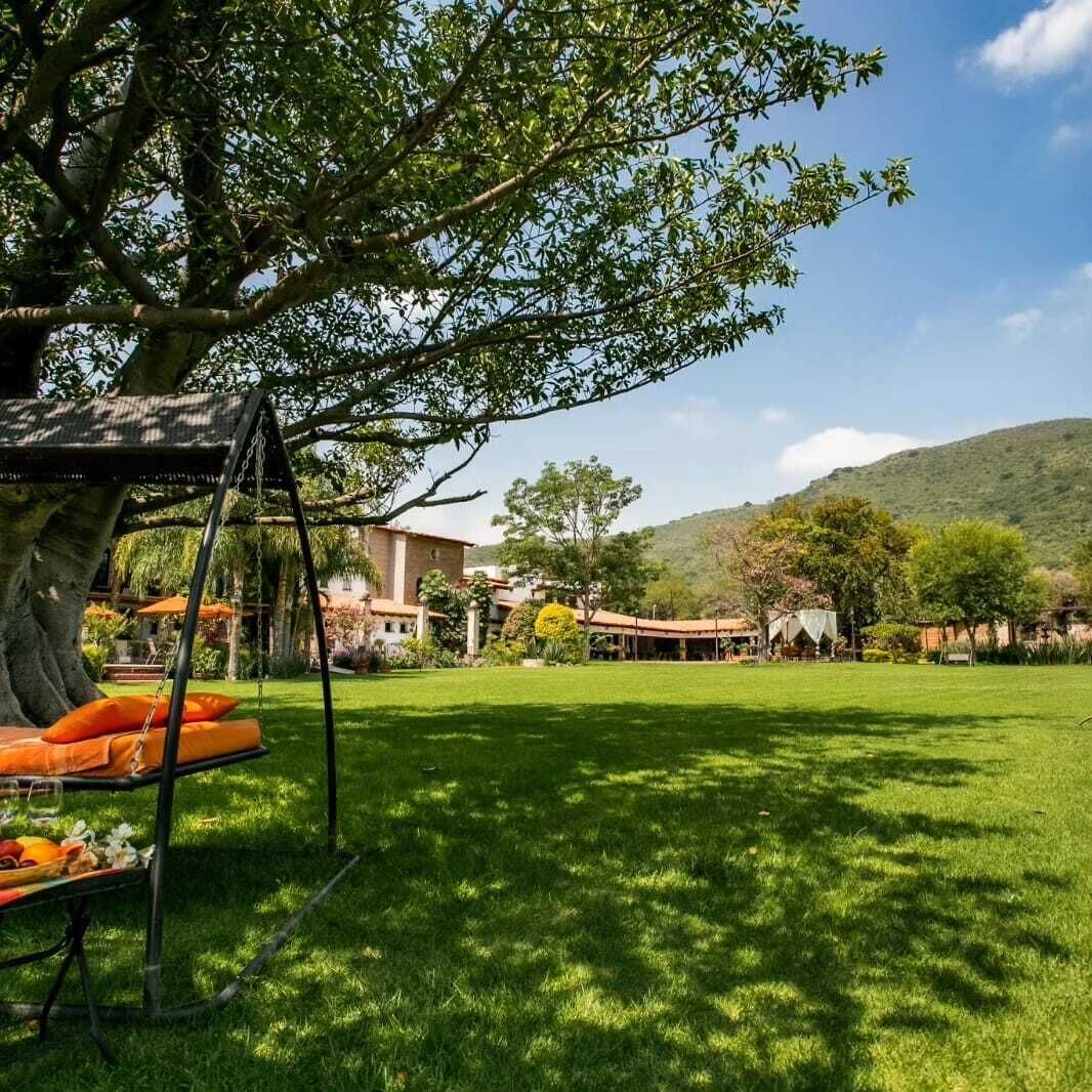 A grassy outdoor area with a swing set, a dining table with orange cushions, and a building with a red roof in the background.