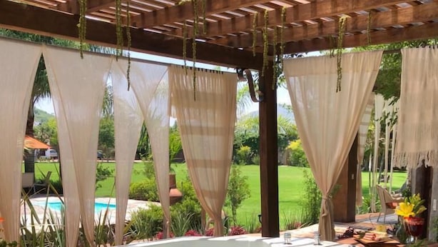 Un bain à remous sous une pergola en bois, agrémenté de plantes suspendues et de rideaux légers, entouré d’une végétation luxuriante et offrant une vue sur une piscine.