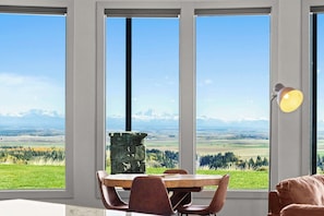 A dining area with a large table and chairs, a stone fireplace, and a view of a mountainous landscape through large windows.