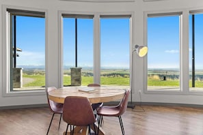 A room with a large round wooden table, four chairs, and a view of a green landscape through large windows.