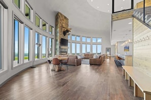 A spacious living area with a fireplace, large windows, and a high ceiling.