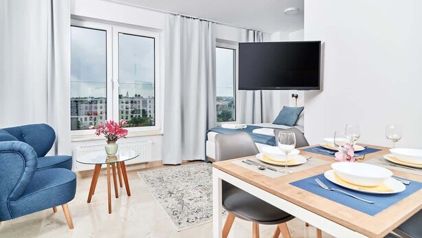 A modern hotel room with a bed, a TV mounted on the wall, a dining table set for two, and large windows with curtains.