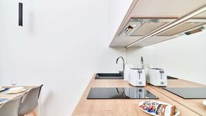 A modern kitchen with a dining table, a black induction cooktop, a kettle, two white toasters, and a plate of snacks.