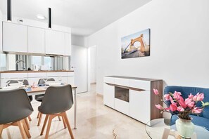 Una cocina moderna con armarios blancos, una zona de comedor con dos sillas, un sofá azul y un jarrón con flores.