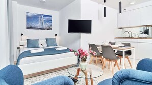 A modern hotel room with a bed, a TV, a dining area, and a kitchen.