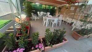 Een terras buiten met witte plastic stoelen en tafels, omringd door potplanten en een bamboomschutting.