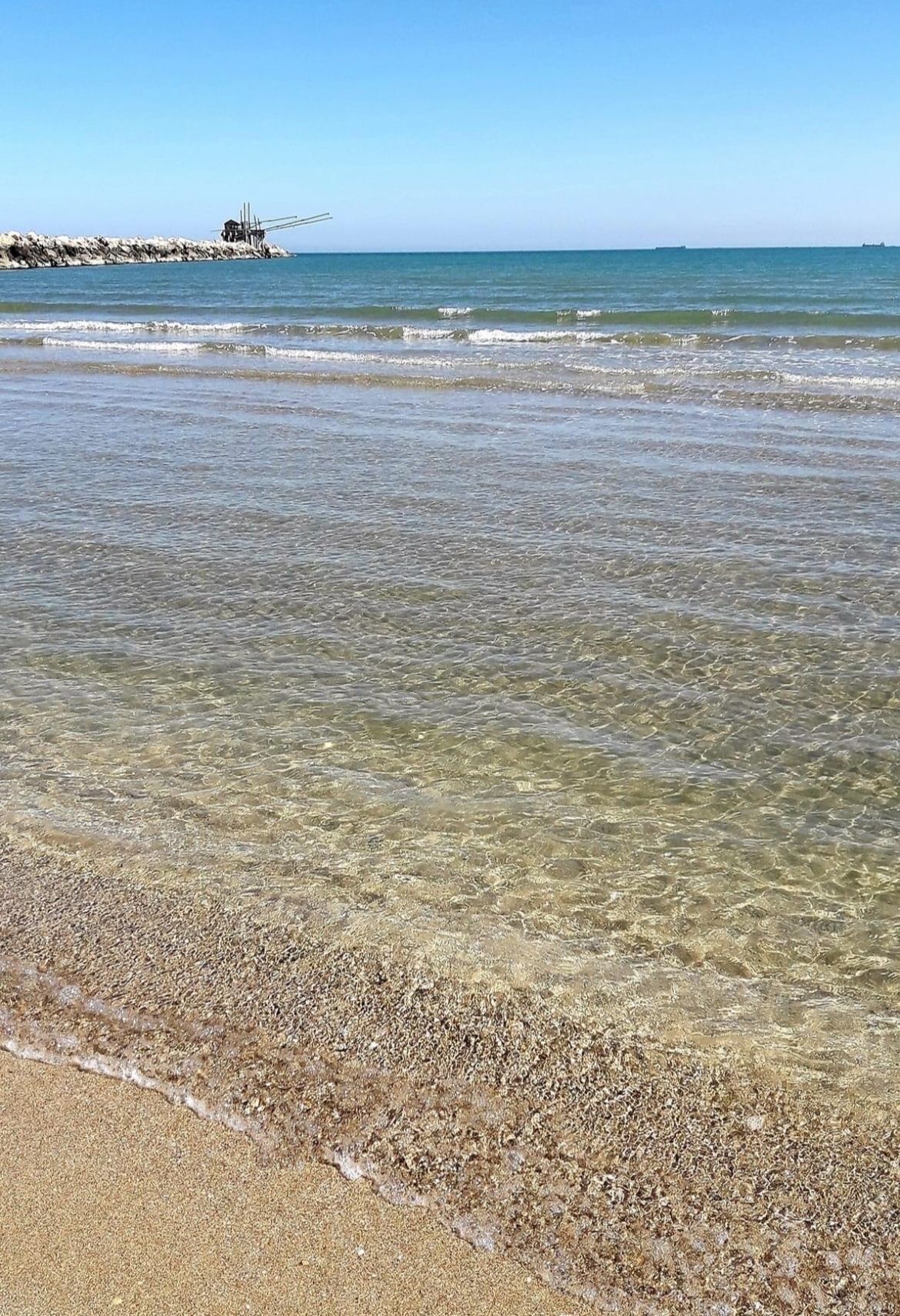 Une plage avec une eau claire et peu profonde, un rivage de galets et un promontoire rocheux visible au loin.