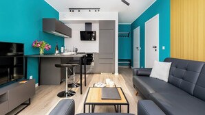 Una sala de estar moderna con un sofá, una mesa de centro y un televisor. Hay una zona de cocina con un refrigerador, estufa y lavaplatos.