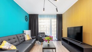 Una sala de estar moderna con un sofá negro, una mesa de centro, un televisor sobre un mueble de madera y un ventanal con cortinas.