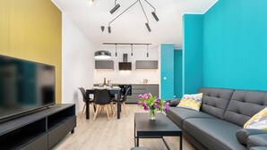 Una sala moderna con un sofá negro, una mesa de centro y un televisor sobre un mueble negro. Hay una zona de comedor con una mesa y sillas, y una cocina al fondo.
