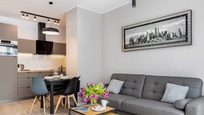 Un salon moderne avec un canapé gris, une table à manger avec des chaises, une petite table basse sur laquelle reposent un livre et un vase de fleurs, et un tableau représentant un paysage urbain accroché au mur.