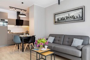 Uma sala de estar moderna com um sofá cinzento, uma mesa de jantar com cadeiras, uma pequena mesa de centro com um livro e flores, e uma pintura de paisagem urbana na parede.