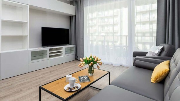 Una sala de estar moderna con un televisor de pantalla plana, un sofá con un cojín amarillo, una mesita de centro con una bandeja de bombones y un jarrón de tulipanes.