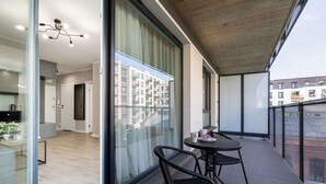Un balcón moderno con puerta de cristal que da a una cocina, una zona de comedor con mesa y sillas, y vistas a edificios residenciales.