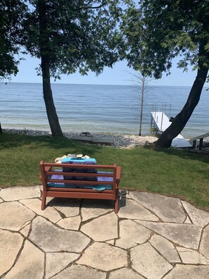 A wooden bench with cushions on a stone patio overlooking a body of water, with trees and a dock in the background. - 3 Bedroom, 2 bath cottage on the shores of Garrett Bay with private beach  (Ellison Bay)