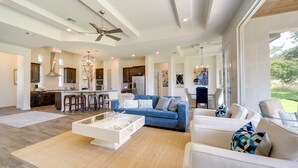 A spacious living area with a blue sofa, white armchairs, and a coffee table. There is a kitchen in the background with modern appliances and a dining area with a chandelier. - 'Cascada at Canyon Lake' ~ 1 Mi to Wedding Venues! (Spring Branch)