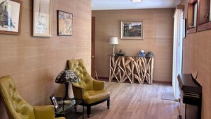Una habitación con piso de madera, un piano, dos sillones, una mesa con lámpara y cuadros enmarcados en las paredes.