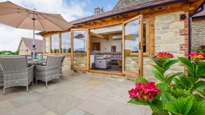 An open-plan living area with a stone exterior, a glass door leading to a dining area with wicker furniture, and a patio with a table and chairs.