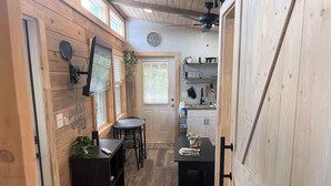 A compact kitchen with wooden walls, a dining area with a table and chairs, and a small black table with items on it. - Lake Side Tiny home in Punta Gorda, Florida (Punta Gorda)