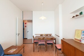 Una habitación con una mesa de comedor, sillas, un armario pequeño, una lámpara colgante y una puerta que da a otra estancia.