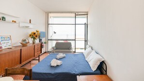 Ein kleiner, gut beleuchteter Raum mit einem Bett, einem Schreibtisch und einem Stuhl. Es gibt ein Fenster mit Jalousien, ein Regal mit Dekoration und einen Blick nach draußen.
