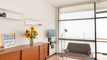 Une pièce avec un meuble en bois, un vase rempli de tournesols, un canapé et une grande fenêtre avec des stores.