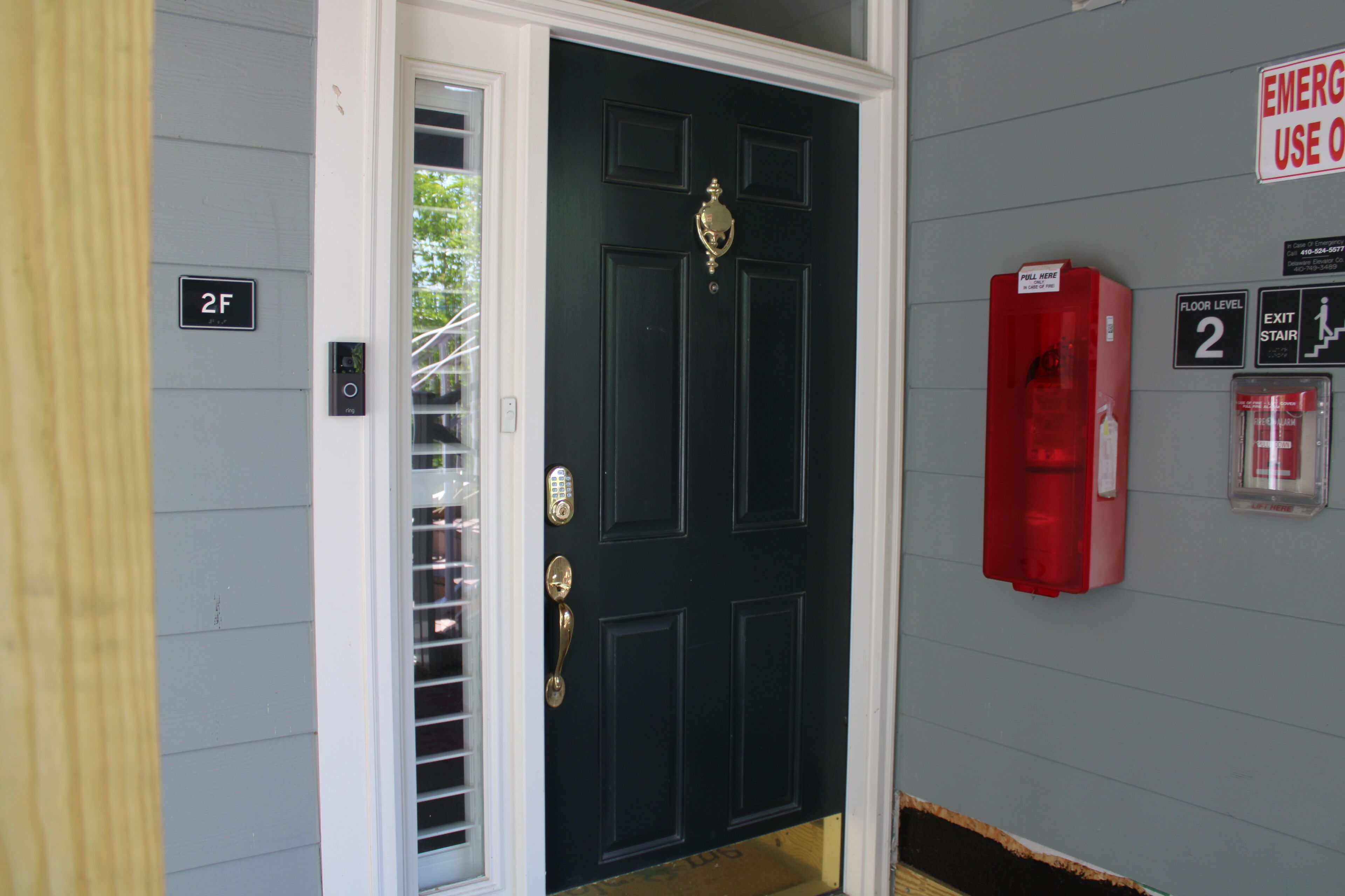 A black door with a brass handle, a fire alarm, and a floor level indicator on a gray wall.